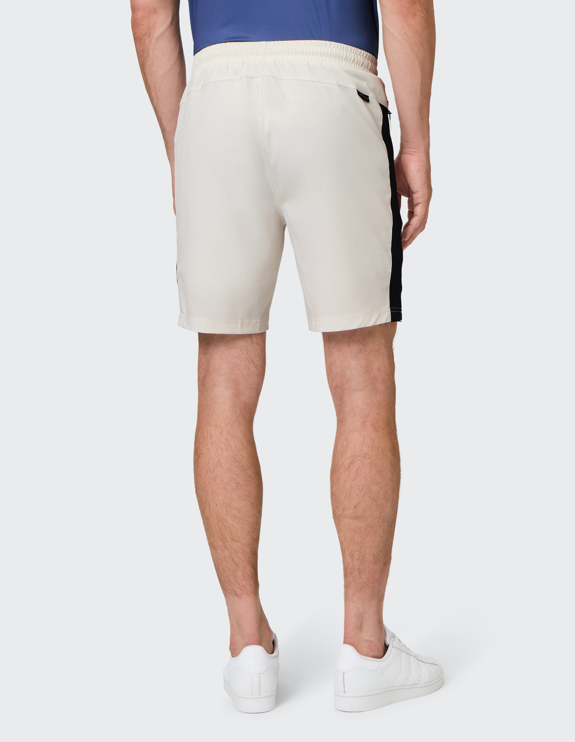 Ein Mann in einem blauen T-Shirt und weißen Shorts mit schwarzen Streifen steht rückwärts, trägt weiße Sneaker vor grauem Hintergrund.