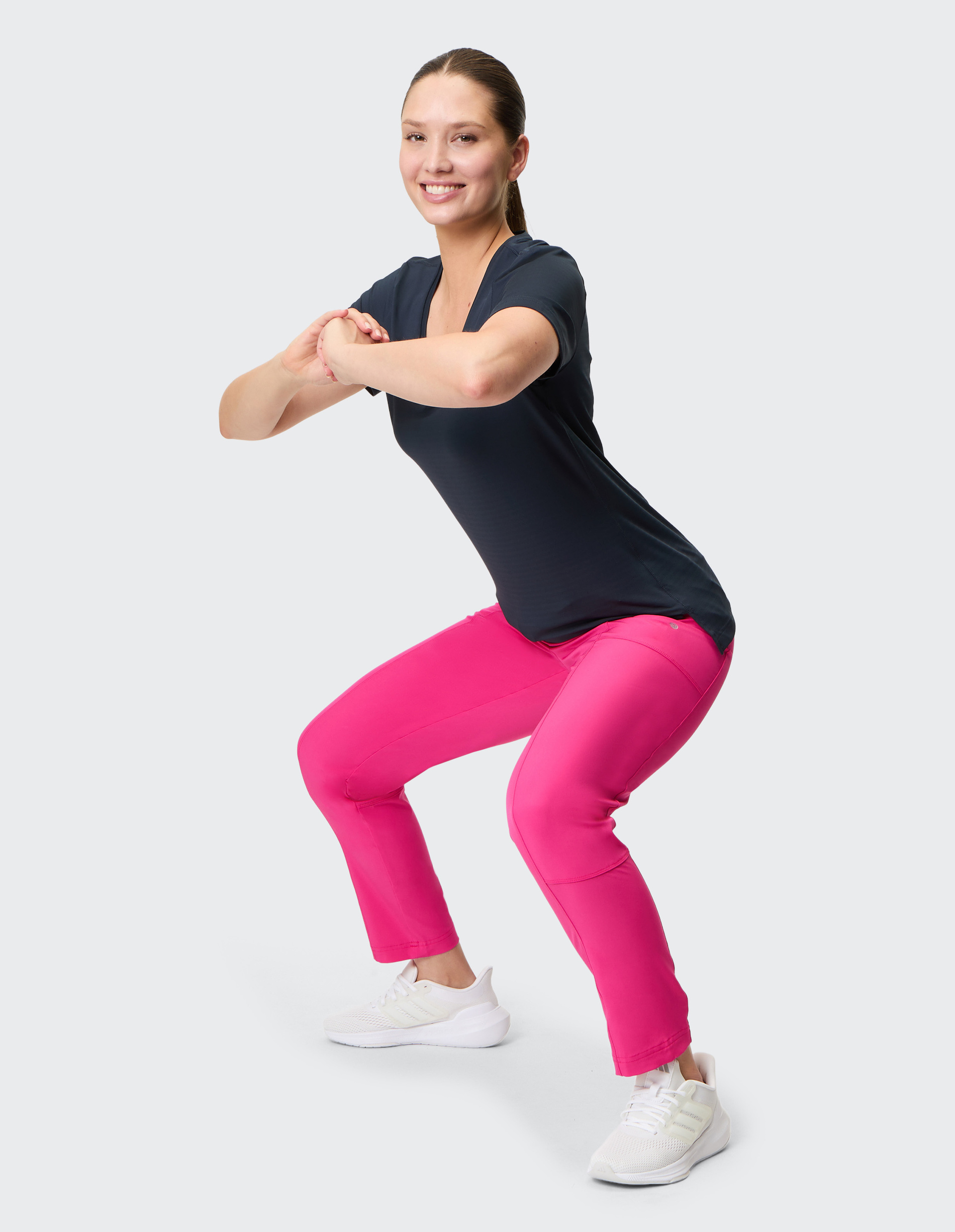 Frau in dunklem T-Shirt und pinker Hose macht fröhlich Kniebeugen, mit weißen Turnschuhen auf hellem Hintergrund, Hände vor der Brust.
